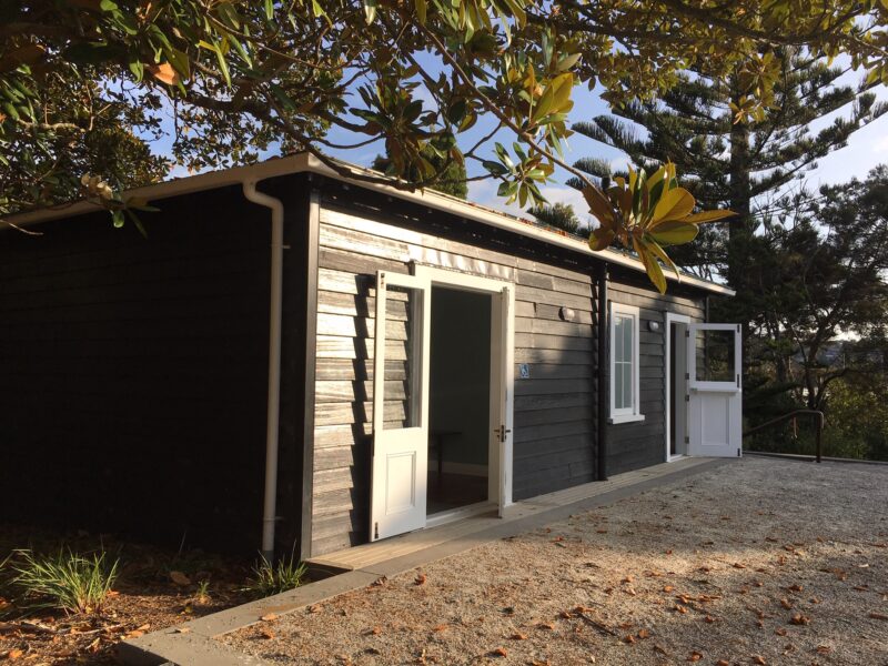 Chichester Cottage: a small dark-stained weatherboard building with white doors standing open onto a gravel forecourt, under a magnolia
