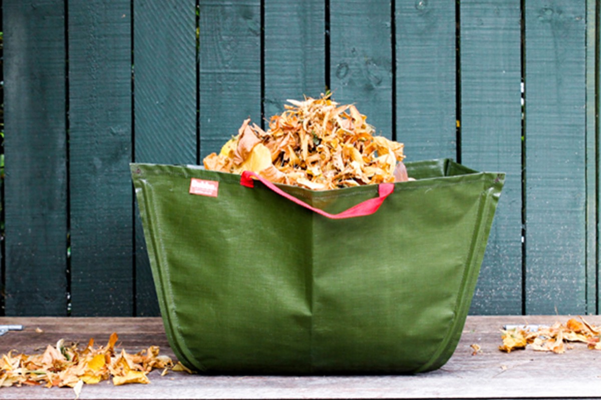 A green garden bag filled with leaves and prunings, standing against a fence