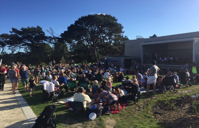 A crowd seated on the grass in front of the amphitheatre stage on a sunny afternoon
