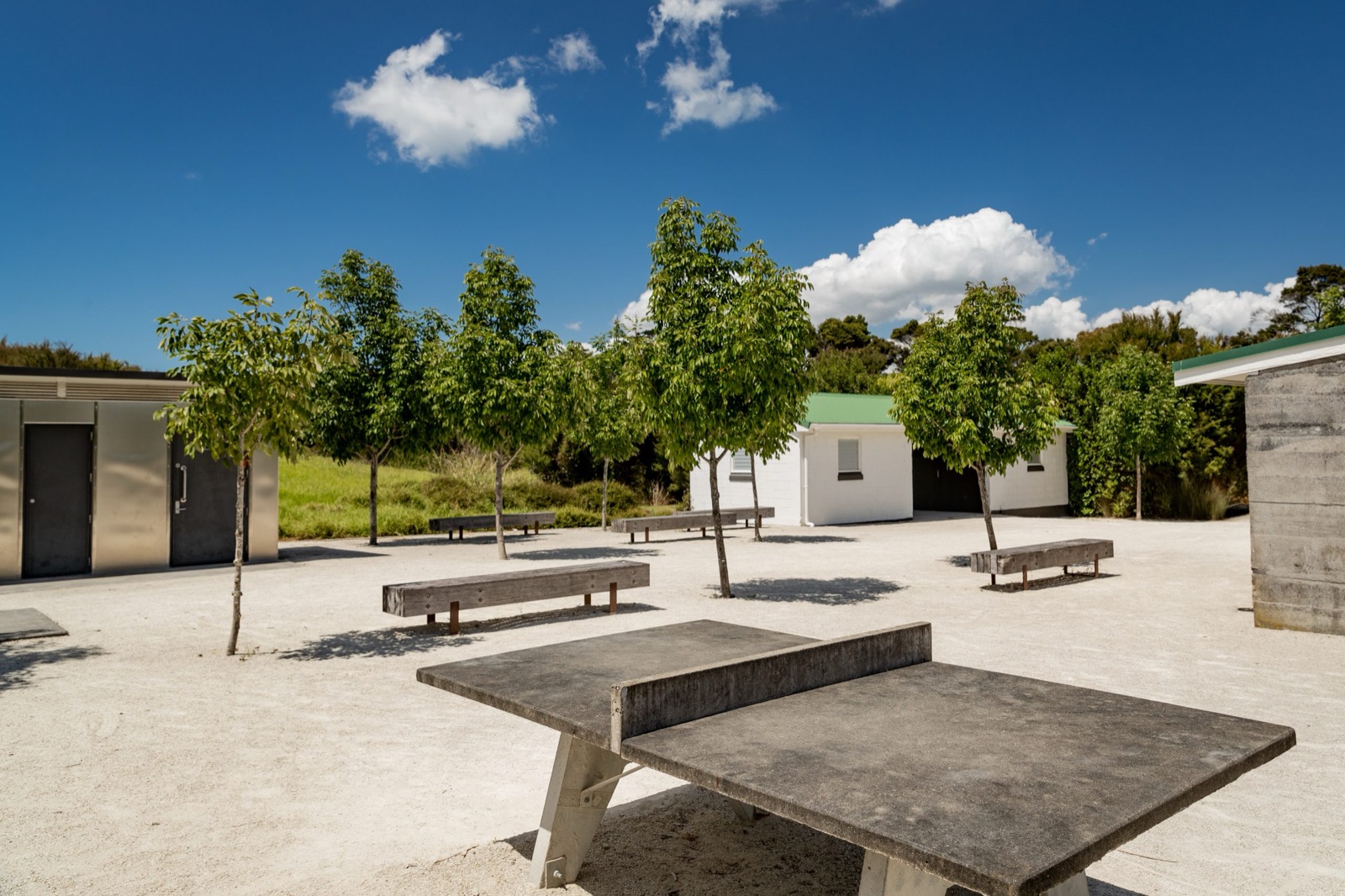 Fixed timber benches and a concrete table-tennis table on a gravel forecourt, with young trees and low buildings around it